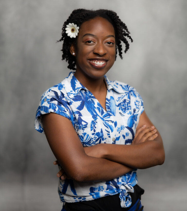 Young woman with a white and blue flowered shirt, with arms crossed and a smile on her face.